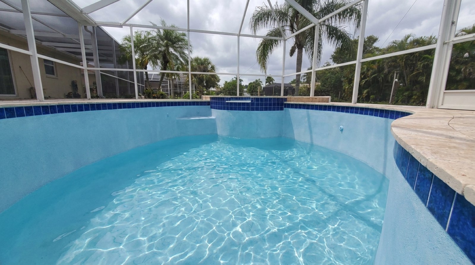 Freshly resurfaced pool with light-blue plaster, blue waterline tile and travertine coping inside a screened enclosure, palms beyond